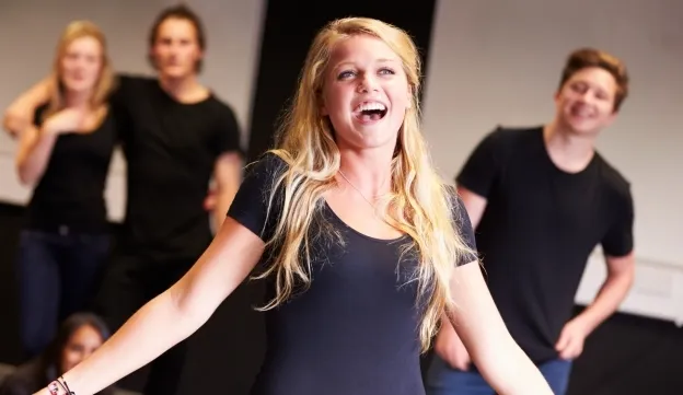 A young woman in a black shirt stands smiling with arms open performing acting